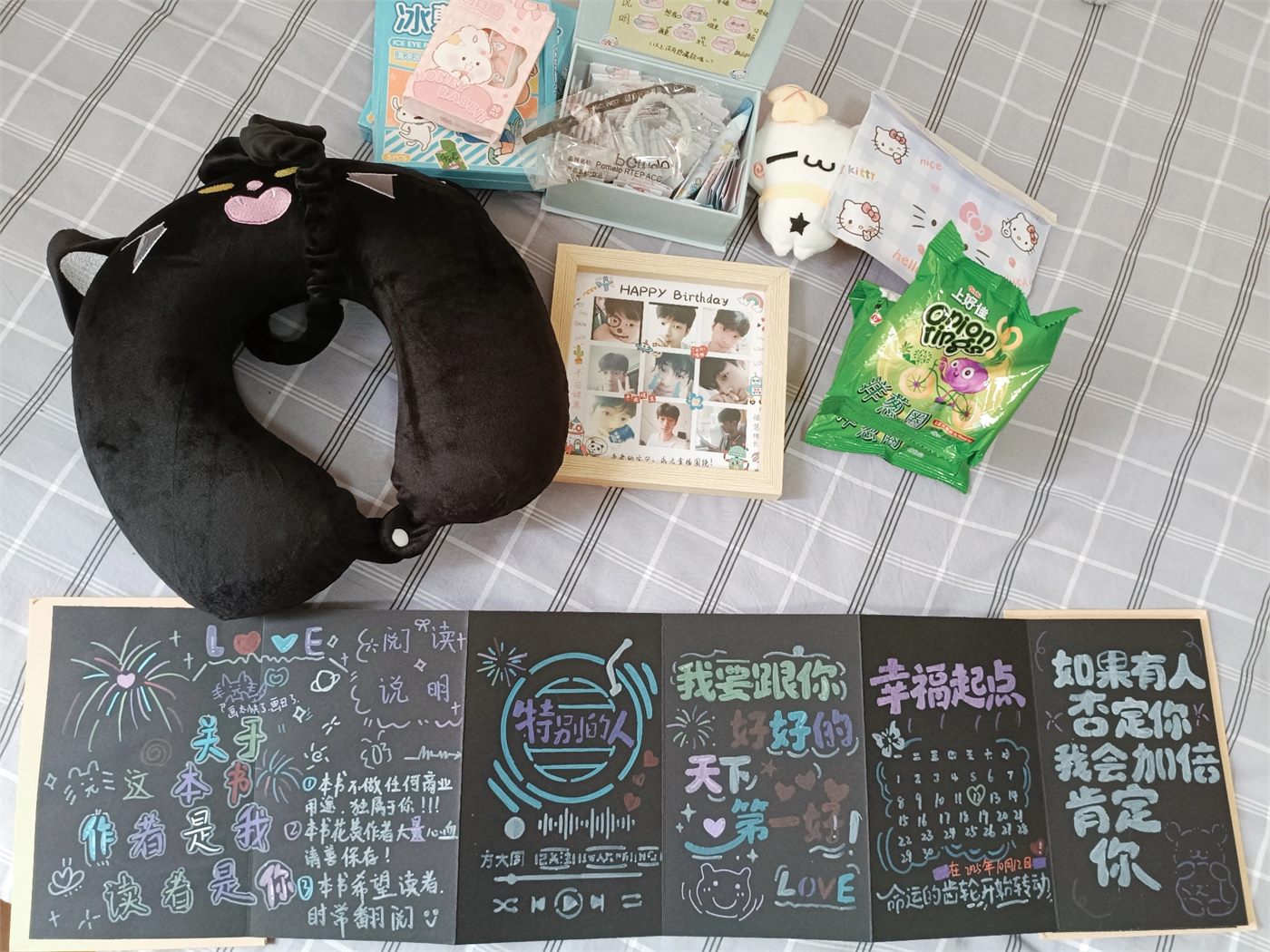 灰色格子床单上的猫咪颈枕、生日相框、零食和黑色手工折页相册